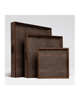 The Amina Tray set by Made Goods features three brown rectangular decorative trays, each with handles and varying in size, arranged in a step formation on a plain white background. Their textured surface and wood trim complement the trays' elegant appearance with a rustic flair.