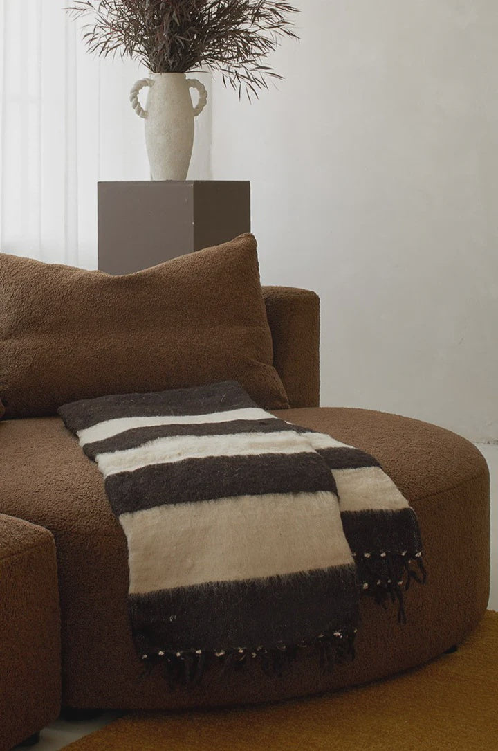 A cozy, brown curved sofa features the elegant Jitana Wholesale Thiago Momo Blanket, with its black and beige stripes offering charm. Behind, a minimalist white vase with dark foliage rests on a brown pedestal. The room's neutral tones and soft lighting echo refined sustainable practices.