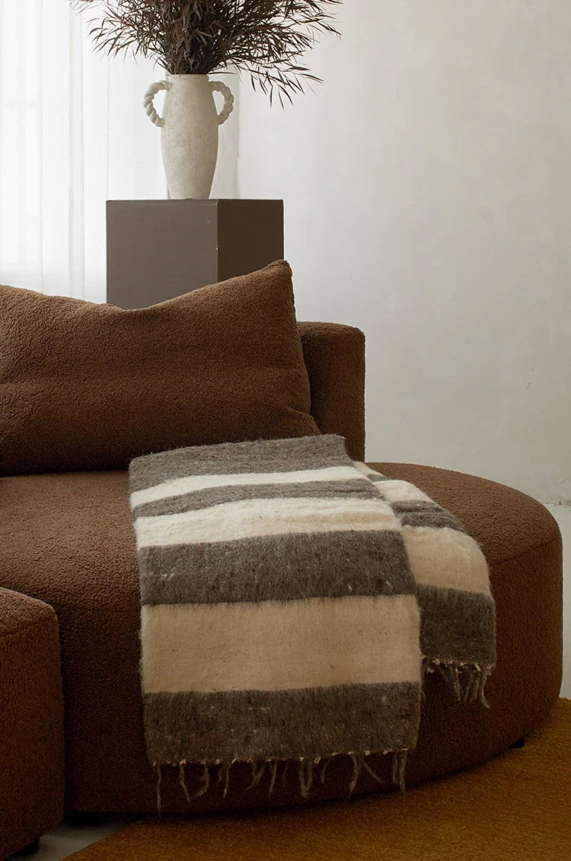 A cozy brown sofa showcases the Thiago Momo Blanket by Jitana Wholesale, expertly crafted by Mayan artisans with dark brown, light brown, and cream stripes. In the background, a tall vase with dried branches sits on a pedestal in a well-lit and inviting setting.