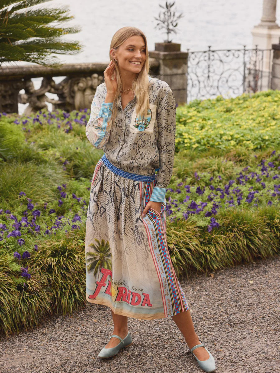 A smiling woman stands on a gravel garden path wearing the Isabel Shirt by Me369 and a colorful "Florida" skirt. She is surrounded by purple flowers, green shrubs, a stone railing, and water in the background.