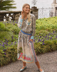 A smiling woman stands on a gravel garden path wearing the Isabel Shirt by Me369 and a colorful "Florida" skirt. She is surrounded by purple flowers, green shrubs, a stone railing, and water in the background.