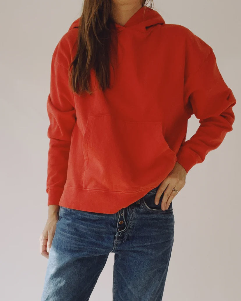 A person wearing &quot;The Hoodie&quot; by Hey Gang, which features a boxy fit in a vibrant shade of red and is crafted from 100% cotton, stands against a plain background. They pair it with blue jeans and have one hand in their pocket while the other rests on their hip. The image cuts off at the shoulders, effectively highlighting the unisex sizing of the hoodie.