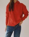 A person wearing "The Hoodie" by Hey Gang, which features a boxy fit in a vibrant shade of red and is crafted from 100% cotton, stands against a plain background. They pair it with blue jeans and have one hand in their pocket while the other rests on their hip. The image cuts off at the shoulders, effectively highlighting the unisex sizing of the hoodie.