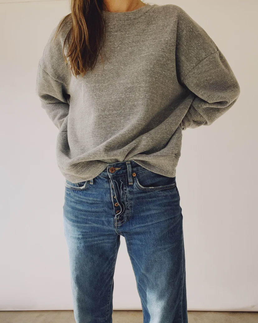 A person is wearing "The Boxy Sweatshirt" from Hey Gang, featuring an oversized vintage fit in a loose gray style, paired with high-waisted blue jeans that have visible buttons. The sweatshirt is slightly tucked into the jeans, and the indoor setting with a plain background emphasizes the unisex appeal of this cozy ensemble.