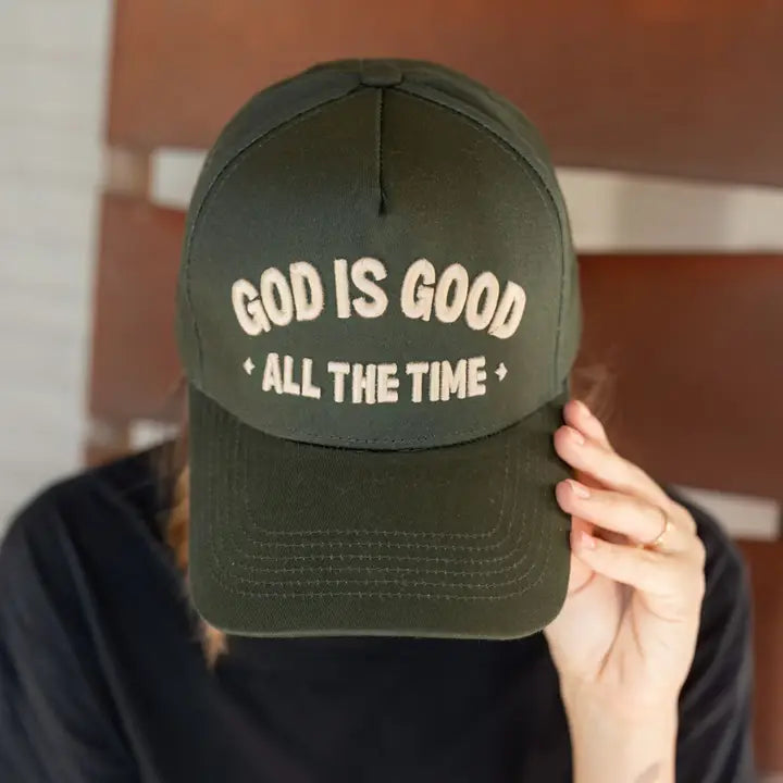 A person with braided hair, partially visible from the front, is wearing a dark green "God is Good Hat" by Faire with the embroidered text "God is good all the time." The person's hand is holding the brim of the hat, slightly covering their face, against a blurred background.