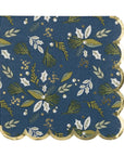 The MME Napkin by Faire is adorned with a blue background showcasing green and white leaves, pine branches, and small berries. It features scalloped edges trimmed in gold.