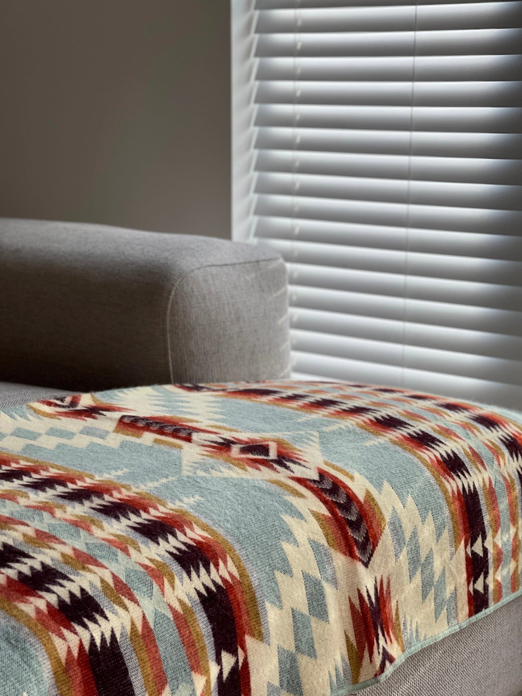 A light gray sofa is draped with the Faire Secoya Throw, crafted by an Ecuadorian artisan. Soft natural light filters through white blinds in the background.