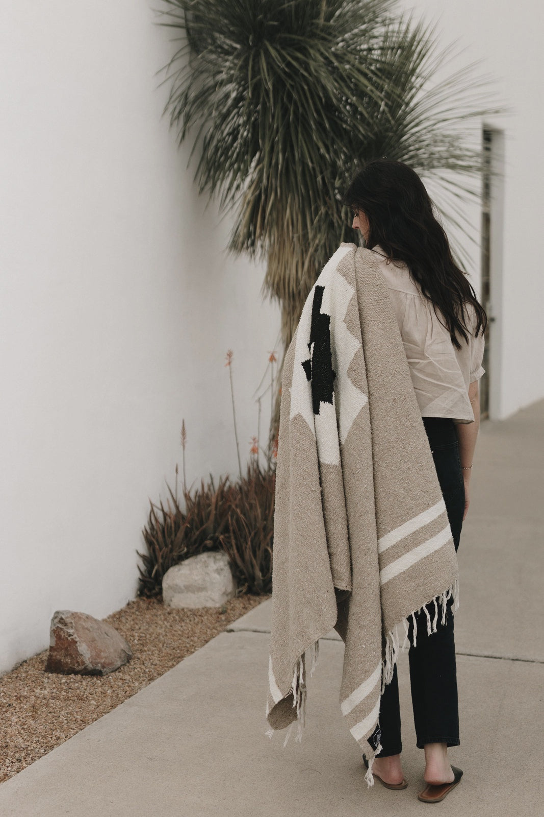 A woman with long dark hair walks beside a white wall and desert plants, draping the Faire TT Handwoven Blanket Taos Two over her shoulder.