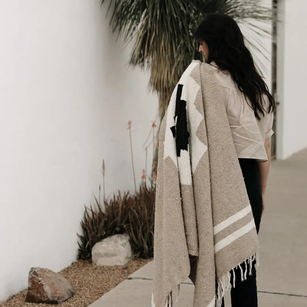 A woman with long dark hair stands outside near a white wall and palm tree, holding the TT Handwoven Blanket by Faire draped over her shoulder. Desert plants and rocks line the walkway beside her.