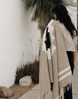 A woman with long dark hair stands outside near a white wall and palm tree, holding the TT Handwoven Blanket by Faire draped over her shoulder. Desert plants and rocks line the walkway beside her.