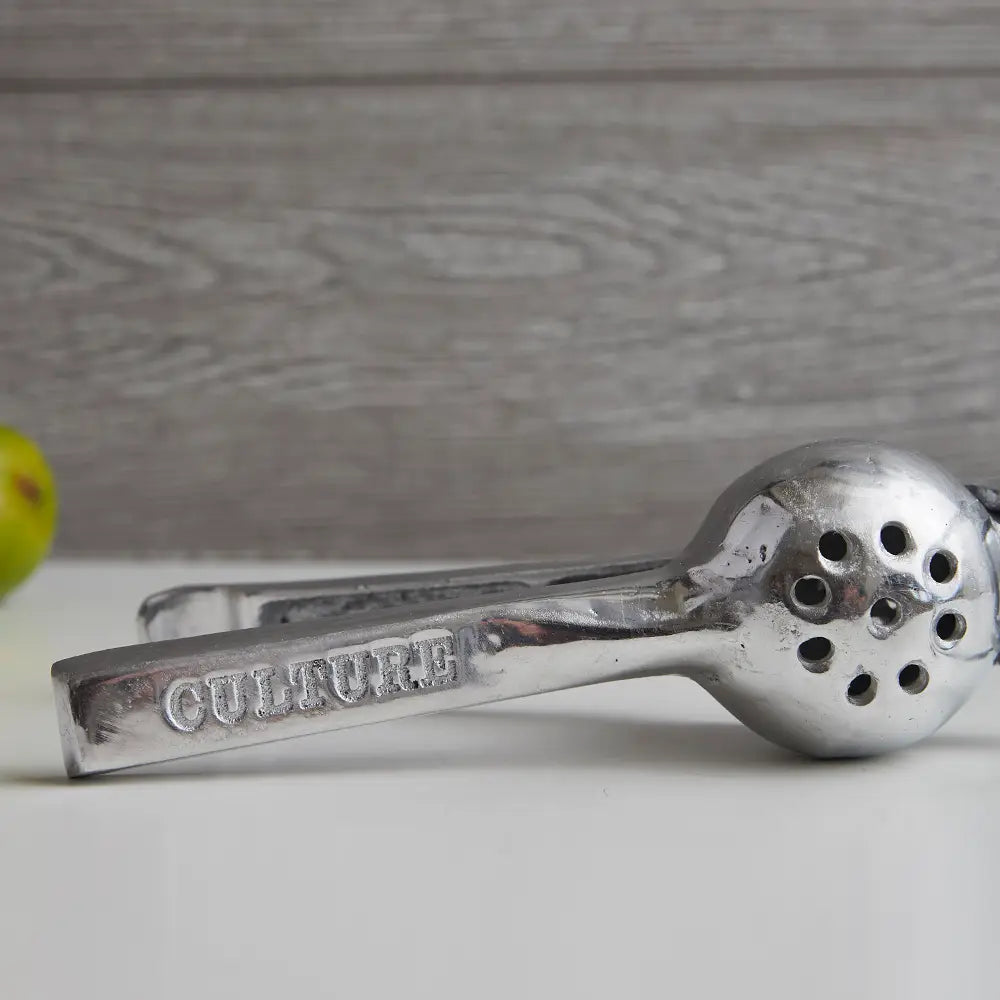 Faire&#39;s Aluminum Hand Juicer, engraved with &quot;CULTURE&quot; on the handle, sits on a white surface against a gray wooden background. A small green fruit is partially visible on the left, ideal for making fresh lemon and lime juice.