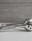 Faire's Aluminum Hand Juicer, engraved with "CULTURE" on the handle, sits on a white surface against a gray wooden background. A small green fruit is partially visible on the left, ideal for making fresh lemon and lime juice.