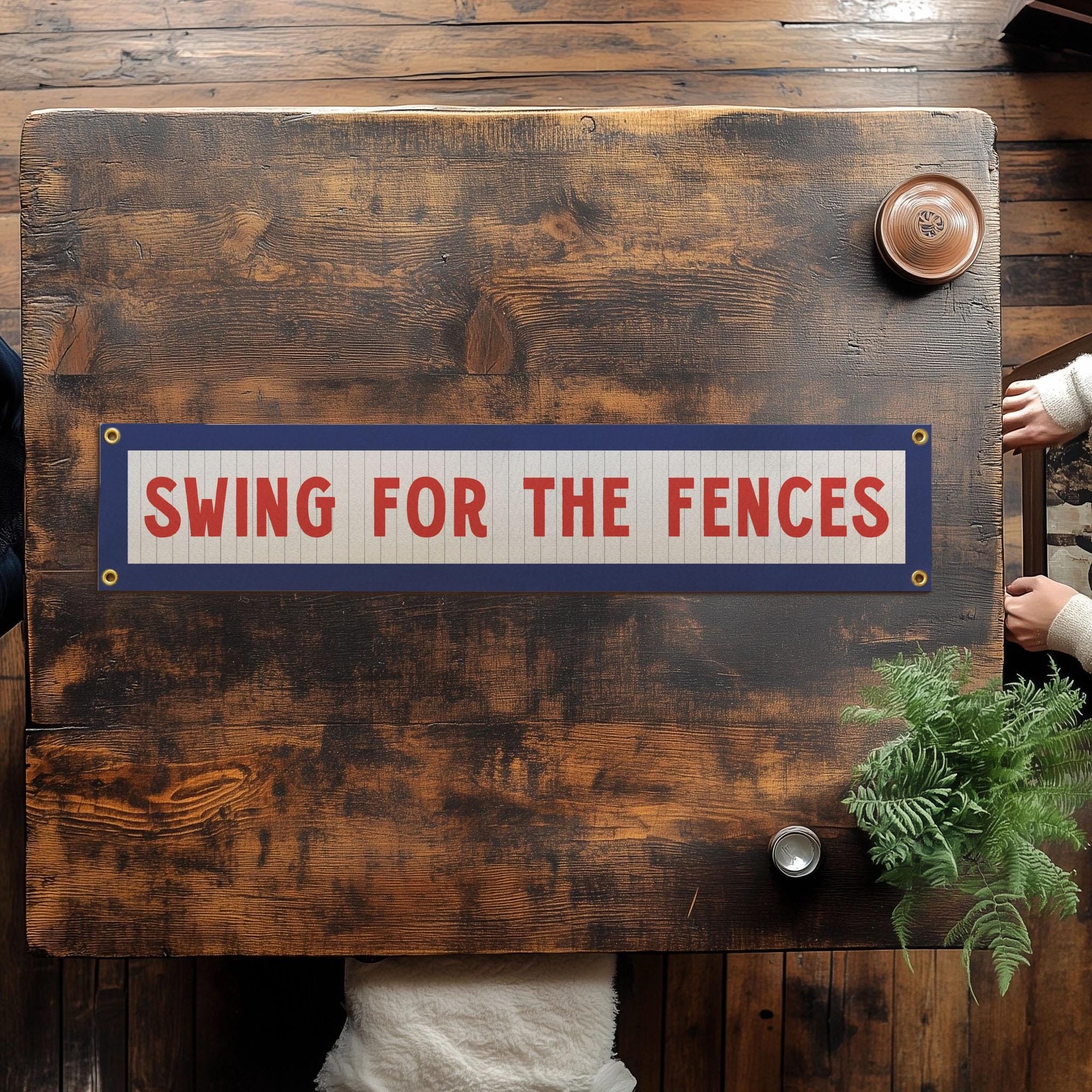 A wooden table features Faire’s Swing for the Fences Banner, a rectangular piece with red lettering. Two people’s hands rest on the table alongside a plant and a cup of coffee.