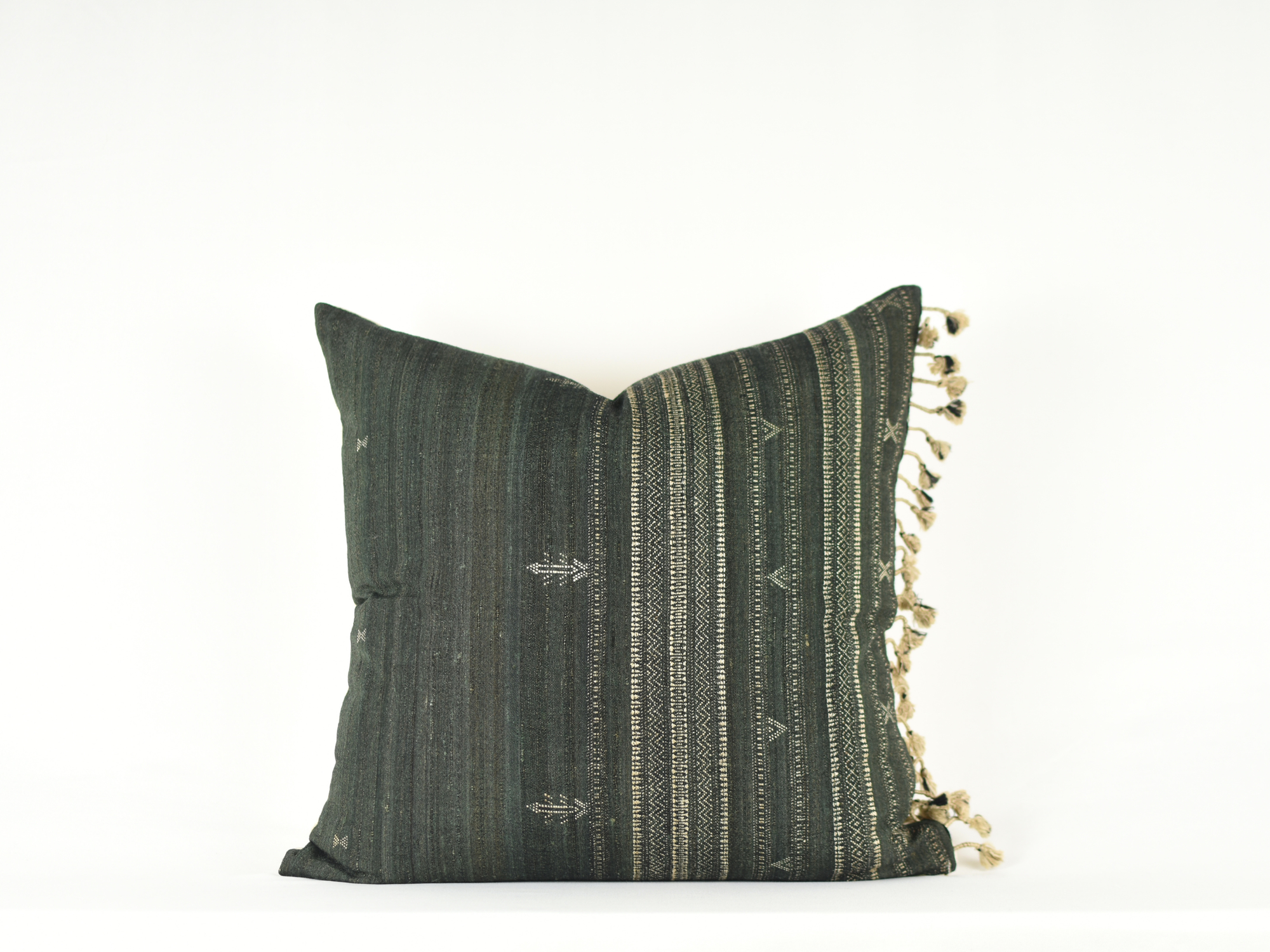 The Dahlia Pom Pillow by Faire is a dark green handmade cover featuring subtle geometric patterns and white tassels on one side, inspired by traditional Gujarat India designs, shown against a plain white background.