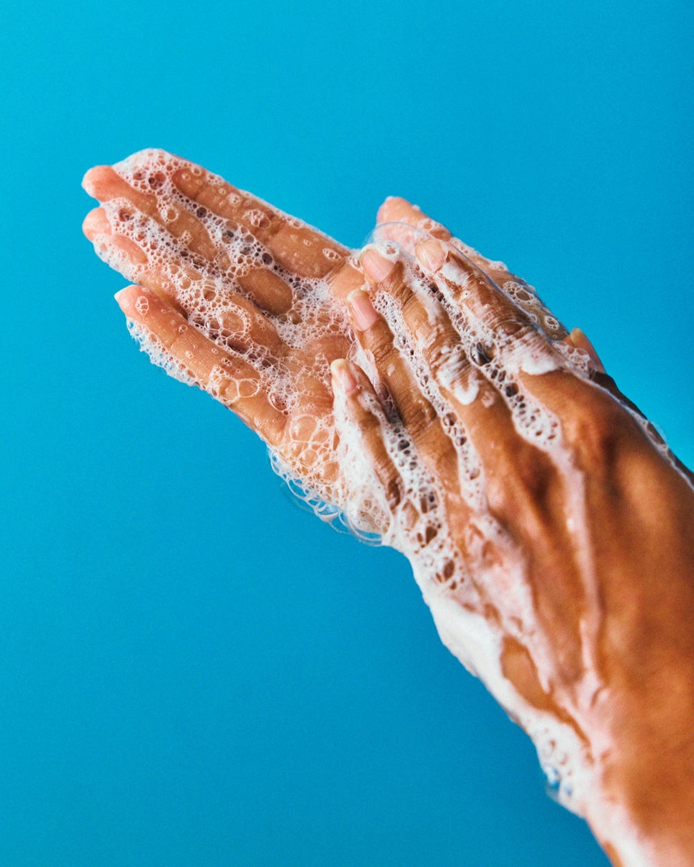 Two hands covered in soapy bubbles are washed together against a bright blue background, showcasing the deep clean and freshness of Faire’s Wild Garden Hand Wash.