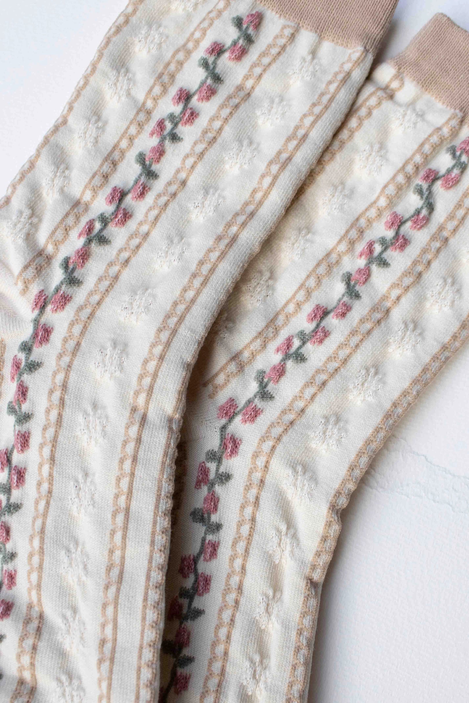 The Faire Vintage Stripe Floral Socks feature a cream knit with beige toes and cuffs, decorated with pink embroidered flowers and green stems along vertical stripes.