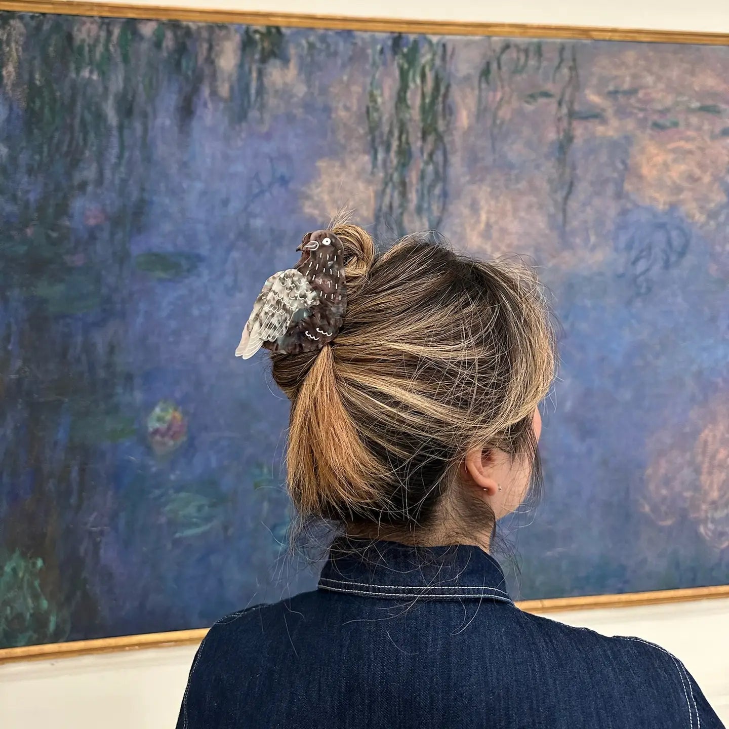 A person with light brown hair in a bun, secured with Faire's Hand Painted Pigeon Clip, stands before an impressionist painting of water lilies in soft blue-purple hues.