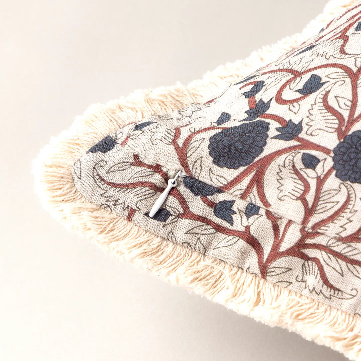 A close-up of Faire's Block Print Pillow shows cream fringe edges, a blue and brown floral pattern, and a small metal zipper pull along the seam of this vintage-style decorative pillow.