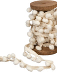 The Faire Christmas Ribbon features cream-colored pom pom trim elegantly wrapped on a wooden spool, with some trim unwound—making it ideal to pair with tinsel garland for charming Christmas decor.