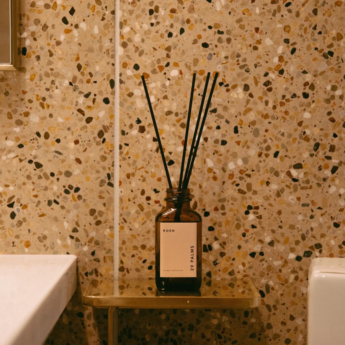 The 29 Palms Reed Diffuser by Faire, with its brown bottle and eucalyptus aroma, sits on a small shelf against a speckled terrazzo bathroom wall. Its minimalistic label enhances the beige and brown palette, adding natural elegance to the scene.