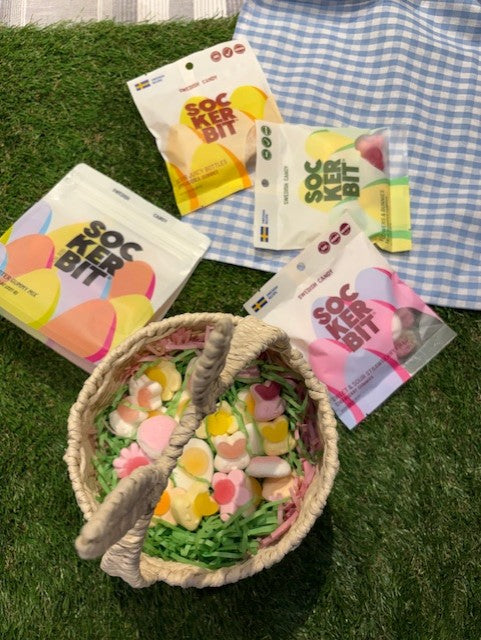 A woven basket with colorful Easter Gummy Mix by Faire sits on artificial grass, next to four packs of non-GMO candy and a blue and white checkered cloth.