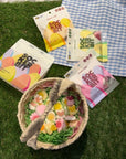 A woven basket with colorful Easter Gummy Mix by Faire sits on artificial grass, next to four packs of non-GMO candy and a blue and white checkered cloth.