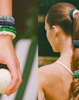 A woman wears Faire's Hair On Point Skinny Scrunchies w/ Pouch in green and blue on her wrist and in her ponytail braid while holding a tennis ball, dressed in green shorts and a white top, outdoors on a tennis court.
