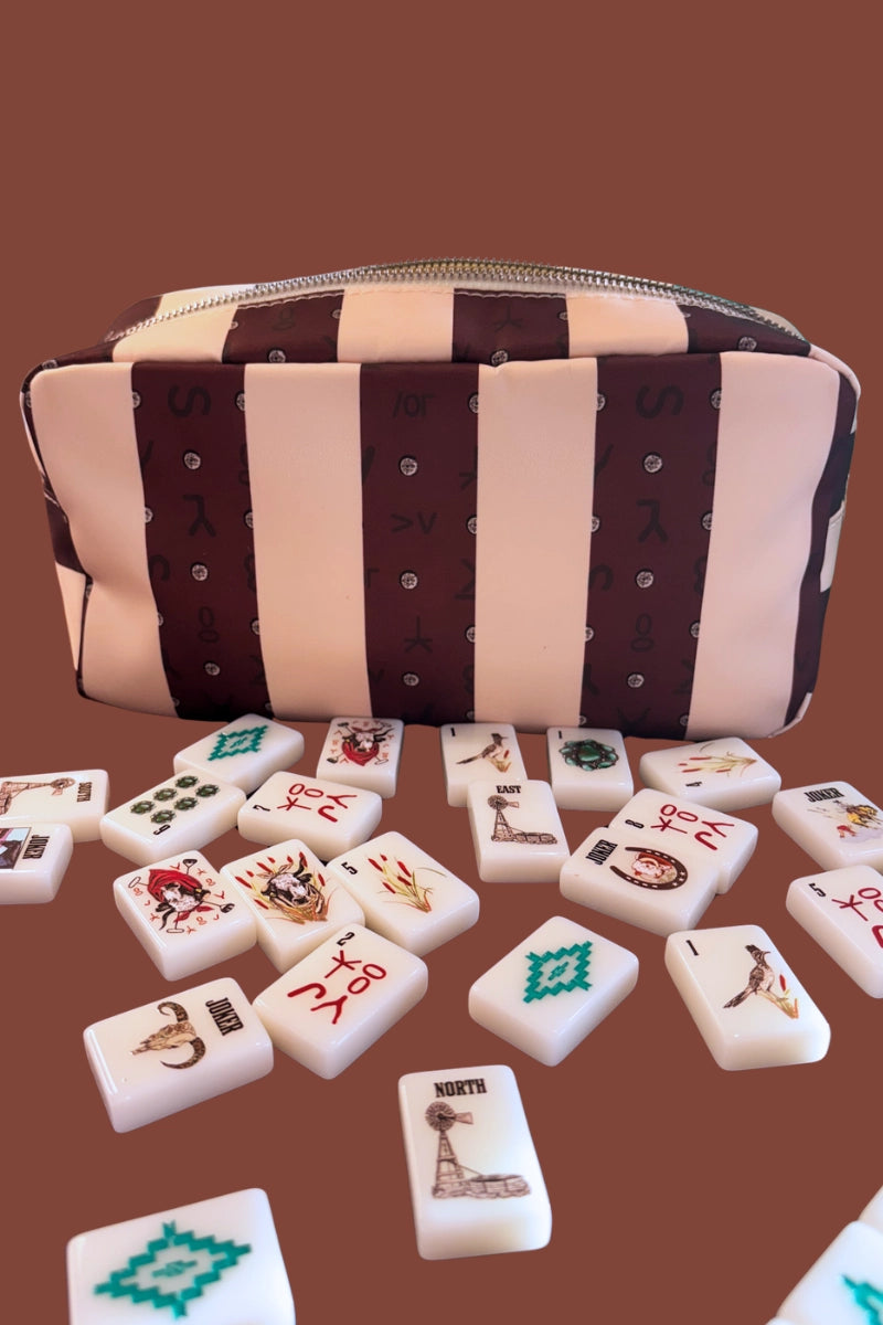 The Faire Wild West Mahjong SET—including tiles, 4 racks, a bag, and mat—is displayed on a brown surface with scattered Western Mahjong tiles showing unique symbols and illustrations.