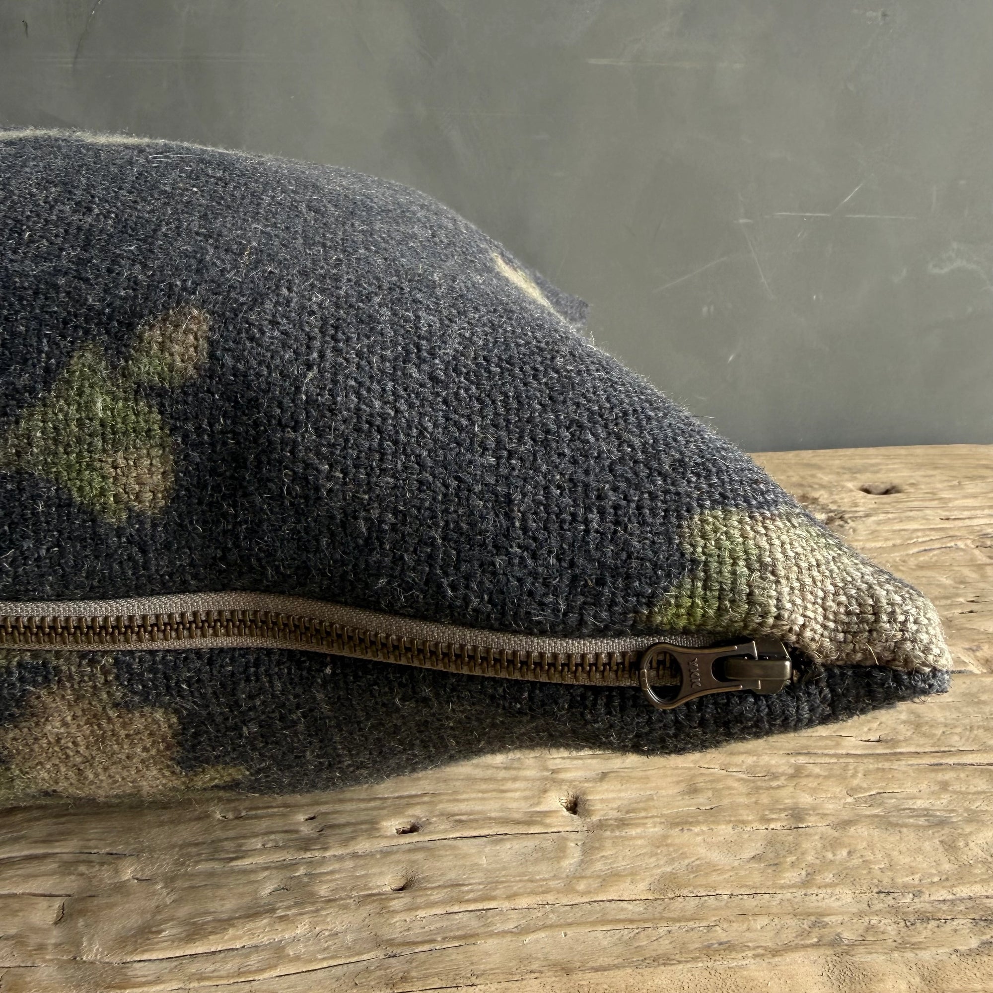 A close-up of Faire's French Linen Roses Pillow, featuring textured dark gray fabric and a partially zipped metal zipper, rests on a rustic wooden surface against a gray wall.