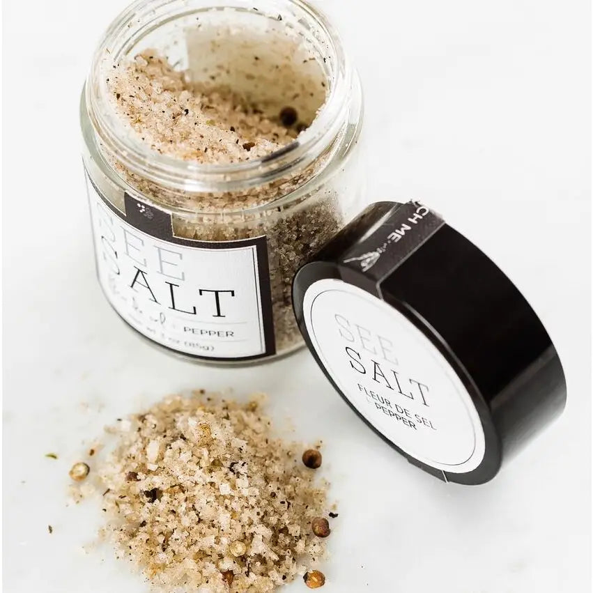 Open glass jar of Fleur de Sel Sea Salt by Faire, with black pepper on the label, sits next to a black lid and scattered salt grains on a white surface.