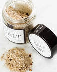 Open glass jar of Fleur de Sel Sea Salt by Faire, with black pepper on the label, sits next to a black lid and scattered salt grains on a white surface.