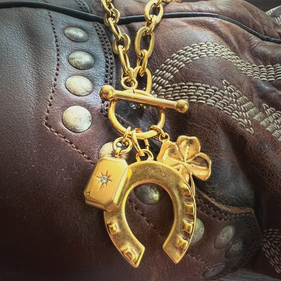 The Lucky Cowgirl Necklace by Faire features a gold-plated horseshoe, shamrock, and hexagonal charm elegantly hanging from a bag with stitched and studded details on a durable stainless steel chain.