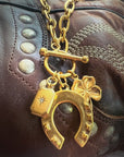 The Lucky Cowgirl Necklace by Faire features a gold-plated horseshoe, shamrock, and hexagonal charm elegantly hanging from a bag with stitched and studded details on a durable stainless steel chain.