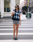 A woman in sunglasses stands on a city crosswalk wearing a white T-shirt, gray shorts, black loafers, and carrying a black bag. She features the White + Warren Mohair Luxe Argyle Vest. Urban buildings and street activity are visible behind her.