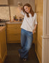 A woman with red hair wears Frank & Eileen’s NYC Trouser and a loose white top, leaning casually against a yellow kitchen cabinet with a smile—epitomizing relaxed New York style.