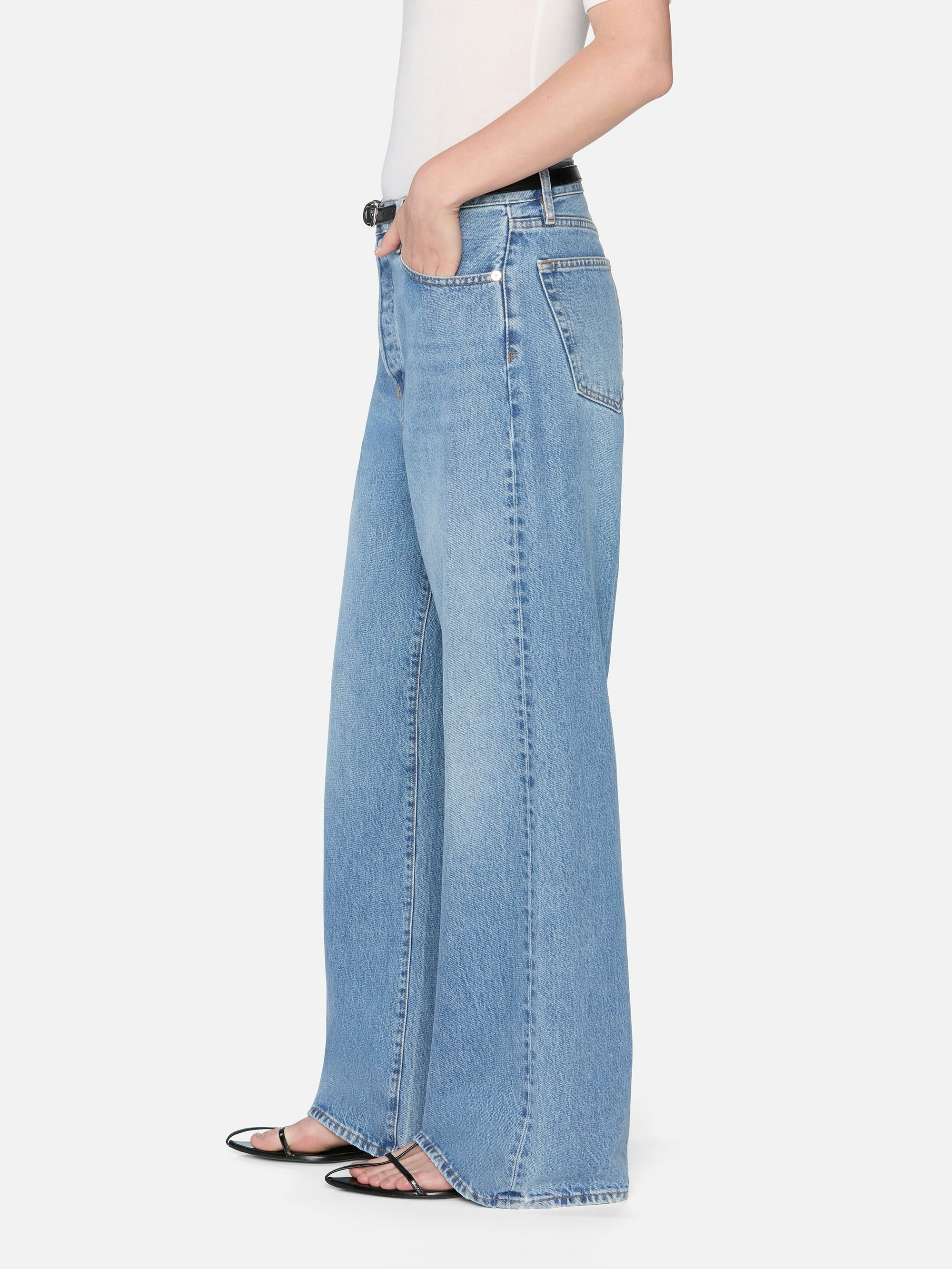 A person wearing FRAME’s The Pixie Loose light blue wide-leg jeans with a petite inseam, a white sleeveless top, and black open-toe sandals stands with one hand in their pocket against a plain white background.