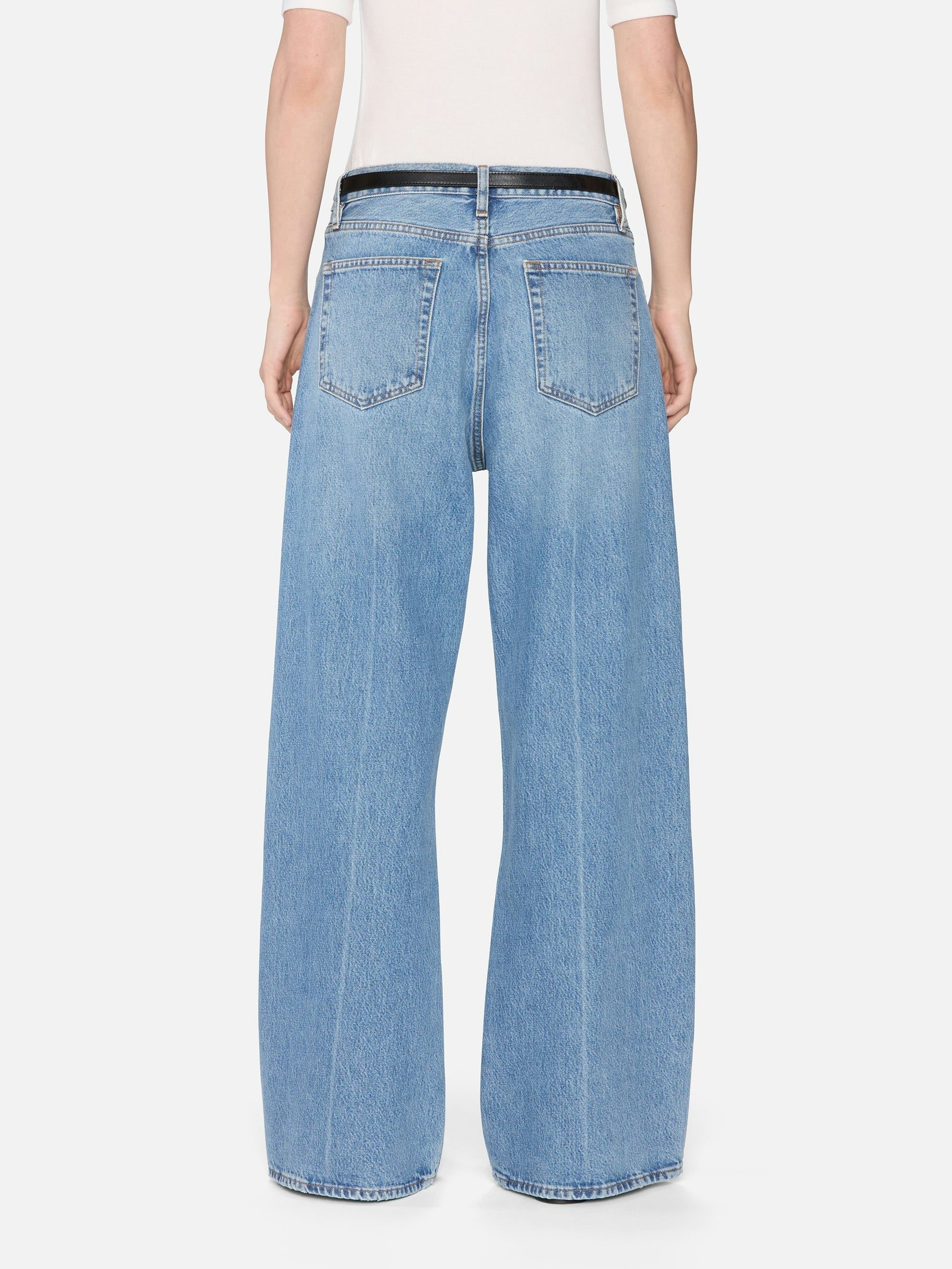A person is seen from behind wearing FRAME's The Pixie Loose jeans—light blue, rigid denim with visible back pockets and a relaxed wide-leg fit. Only the lower back and legs are visible; the jeans are paired with a white top.