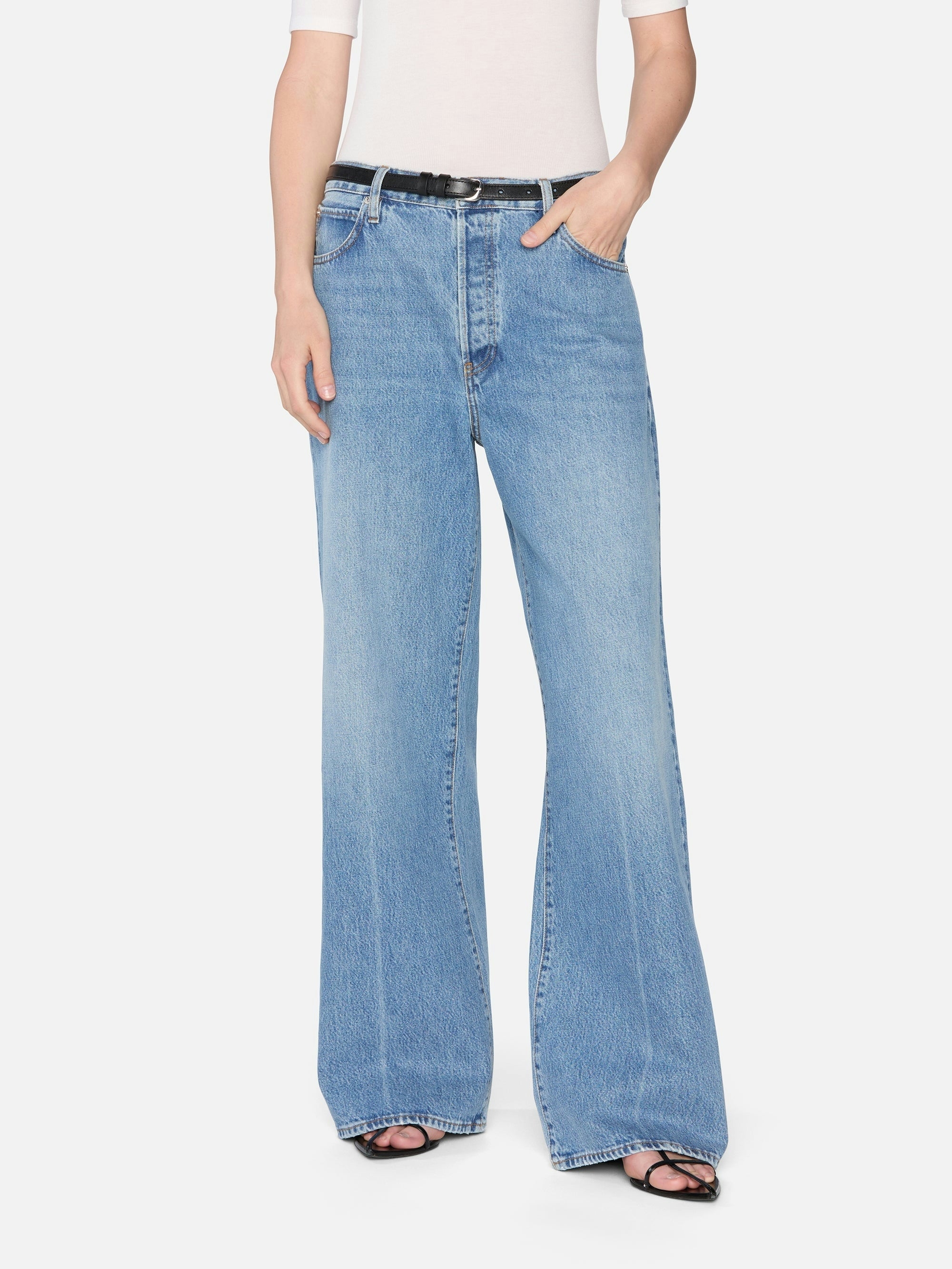 Wearing FRAME's The Pixie Loose light blue, high-waisted wide-leg jeans in rigid denim with a white short-sleeve top, black belt, and black open-toe sandals, a person stands against a plain white background.