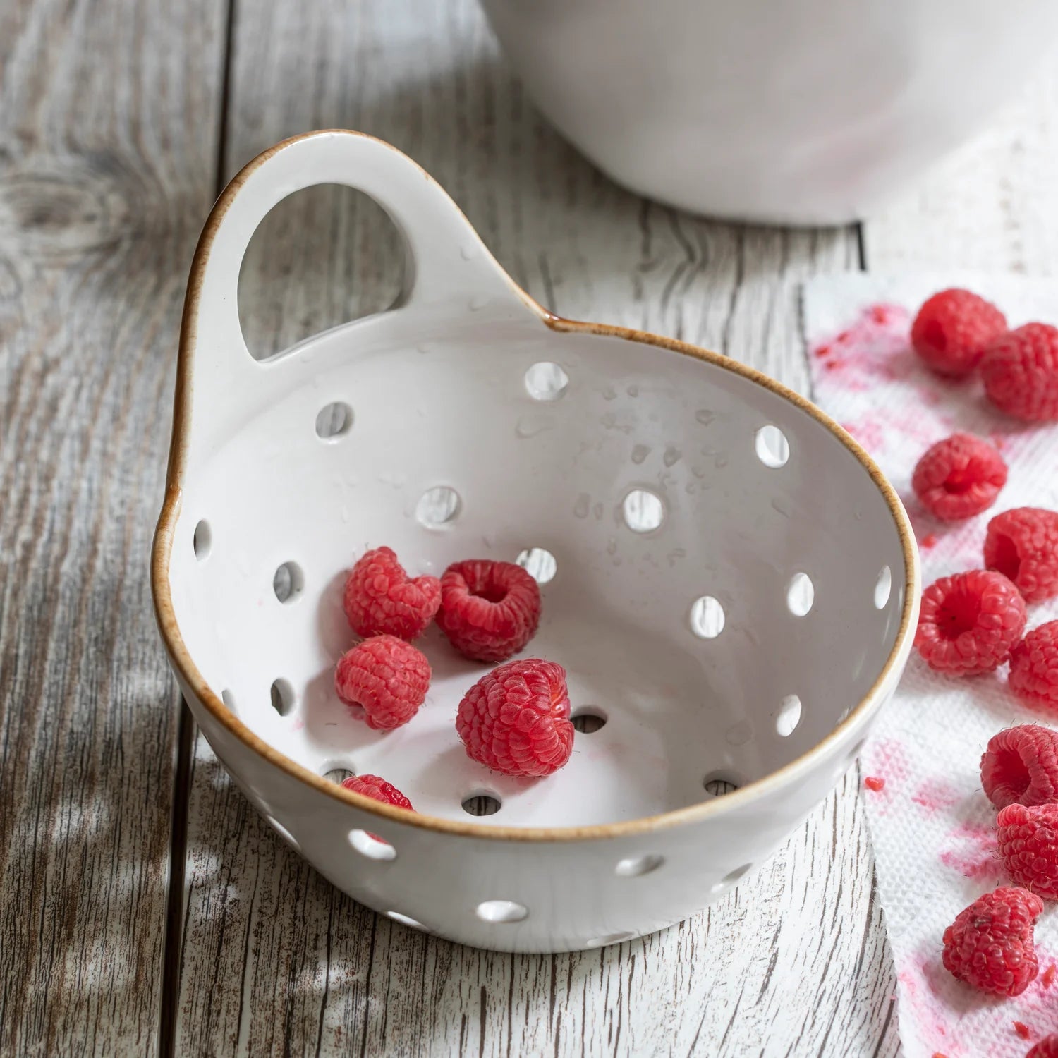 The Yara Colander Basket by Be Home, a handcrafted stoneware piece with a handle, holds fresh raspberries. More raspberries and a white cloth with pink stains sit on the rustic wooden surface, showcasing skilled woman artisanship.