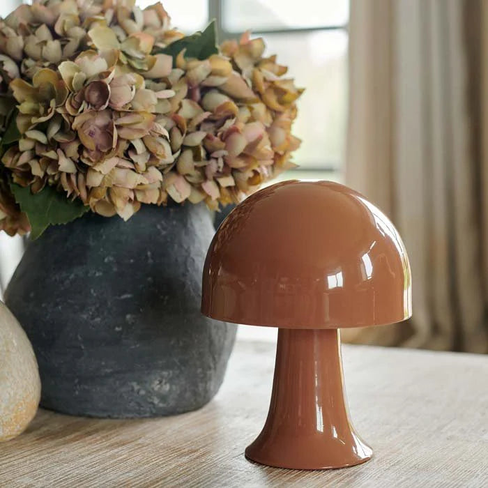 The Julio Portable Table Lamp in Sepia by Abigail Ahern, shaped like a glossy mushroom with an LED bulb, rests on a wooden table. In the background, a large rustic vase holds dried hydrangeas while soft natural light filters through the window.