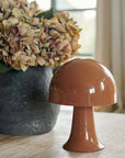 The Julio Portable Table Lamp in Sepia by Abigail Ahern, shaped like a glossy mushroom with an LED bulb, rests on a wooden table. In the background, a large rustic vase holds dried hydrangeas while soft natural light filters through the window.