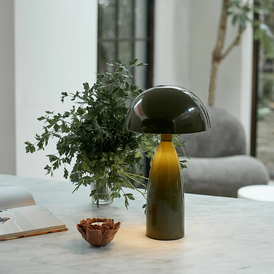The Leoma Lamp Olive by Abigail Ahern sits on a marble surface beside a vase of greenery, a small decorative bowl, and an open book. In the background, there’s a gray chair and a large window.