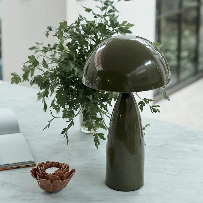 The Leoma Lamp Olive by Abigail Ahern, with its glossy dark green dome shade, is displayed on a marble table alongside a brown ceramic candle holder, an open book, and a glass vase filled with leafy branches in a bright room.
