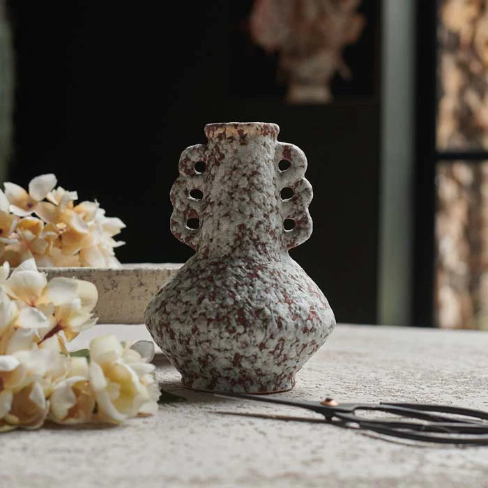 A small, speckled Abigail Ahern Marilla Vase with decorative handles sits on a textured surface beside cream flowers and scissors, set against a soft, blurred background.
