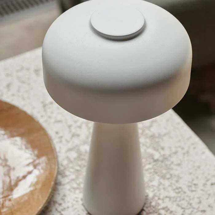 The Abigail Ahern Adelaide LED Lamp White, a modern minimalist table lamp with a round flat shade and conical base, sits on a textured surface near a wooden plate. It's dimmable, portable, and USB rechargeable for convenience.