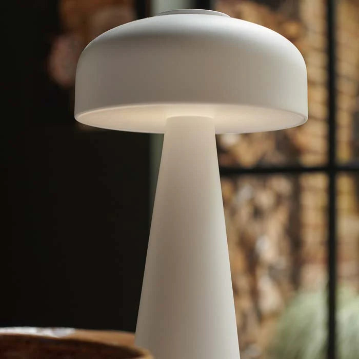 The Adelaide LED Lamp White by Abigail Ahern, a modern mushroom-shaped portable table lamp, glows softly in a warmly lit room with blurred natural elements and a window in the background.
