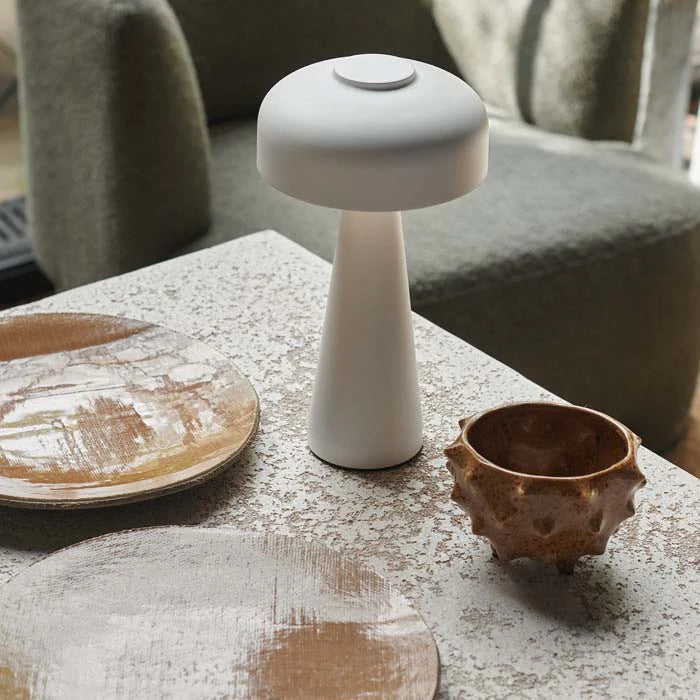 The Adelaide LED Lamp White by Abigail Ahern sits on a textured stone table near two rustic wooden plates and a brown ceramic cup, with a green upholstered chair in the background.