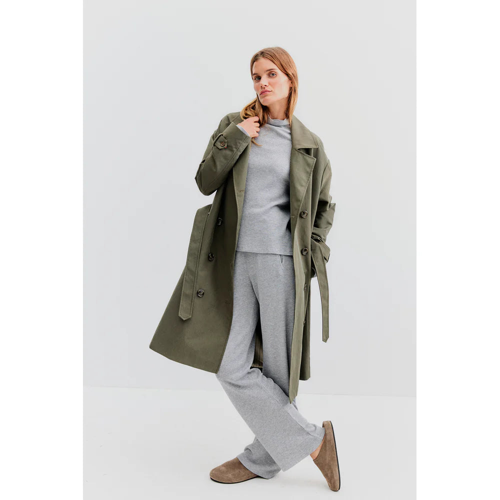 A person poses in an oversized fit, featuring The Rox trench by Kule in chic olive green over a light gray sweater and pants. They wear brown slippers and stand against a plain white backdrop, creating a minimalist and modern look.