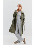 A person poses in an oversized fit, featuring The Rox trench by Kule in chic olive green over a light gray sweater and pants. They wear brown slippers and stand against a plain white backdrop, creating a minimalist and modern look.