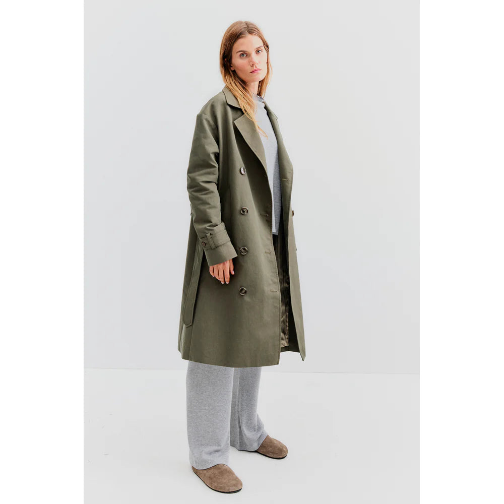 A person poses against a plain background, donned in an oversized fit that includes the chic Kule's The Rox trench coat made from water-resistant fabric in olive green. It's stylishly layered over a grey sweater and wide-leg pants, complemented by brown shoes to complete the look.